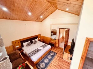 A bedroom with a wooden ceiling, a bed with patterned bedding, a small table, and a chair. - The Rohtang Chalet Cottage (Manali)