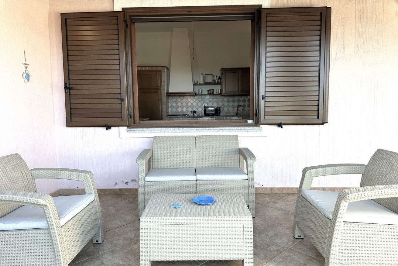 Un espace patio aménagé avec des meubles en osier, une vue sur une cuisine à travers une fenêtre et un sol carrelé.