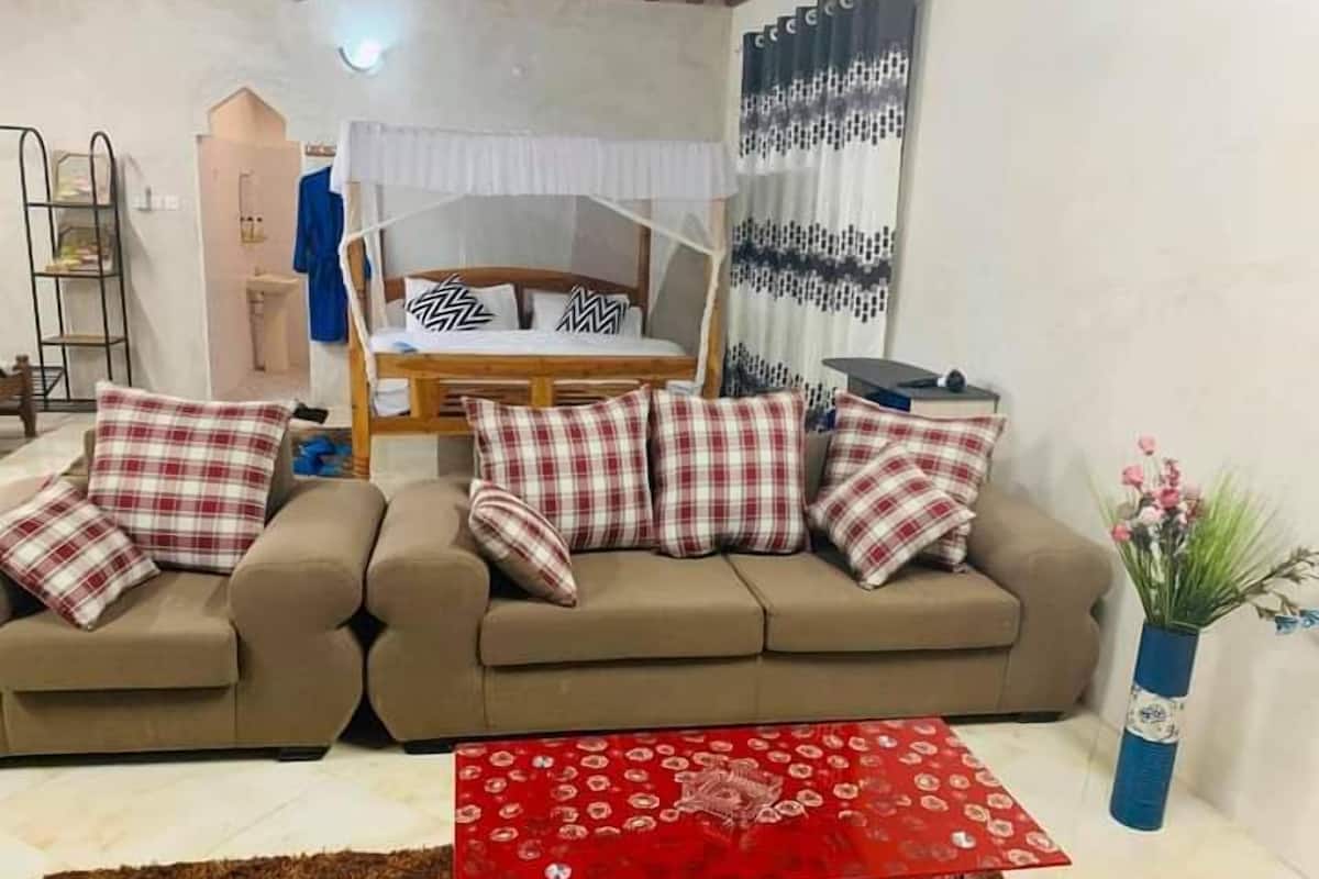 A living room with two sofas, a red coffee table, a bed with a canopy, and a wall with patterned curtains.