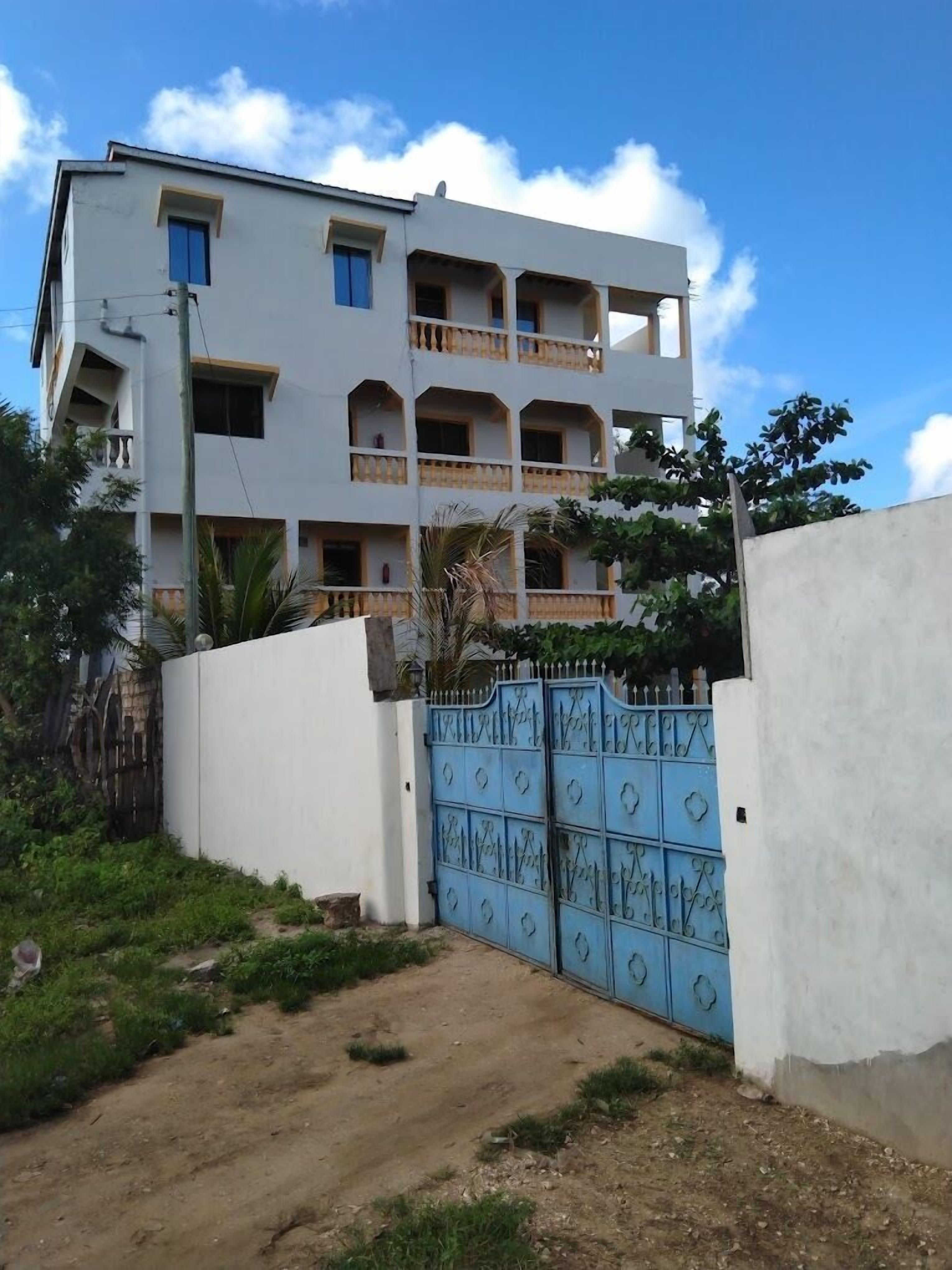 Un edificio de varios pisos con balcones, una entrada con reja y un sendero de tierra que conduce a la puerta.