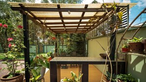 Een overdekt terras met een tafel en stoelen, omgeven door potplanten en een houten pergola.