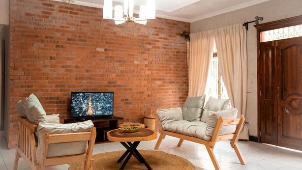 Een woonkamer met een bakstenen muur, een televisie, twee houten stoelen, een ronde salontafel en een houten deur.