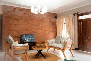A living room with a brick wall, a television, two wooden chairs, a round coffee table, and a wooden door.