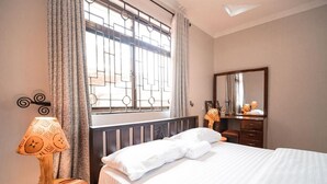 Een net opgemaakt bed met witte lakens, een houten nachtkastje met een lamp met een patroon, een dressoir met spiegel, en een raam met metalen tralies en gordijnen.