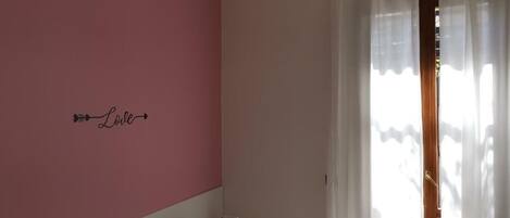 A bedroom with a bed, a pink wall, and a window with sheer curtains.