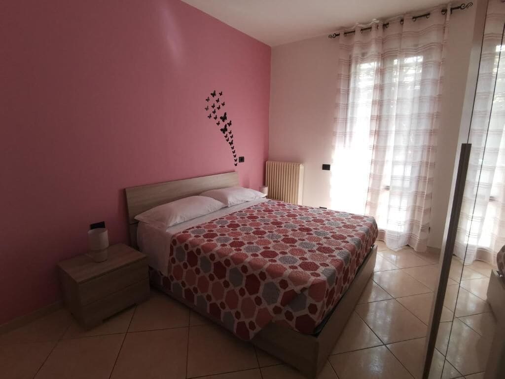 A bedroom with a pink wall, a bed with a patterned bedspread, a nightstand, and a window with sheer curtains.