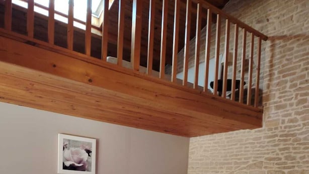 Ein Schlafzimmer mit Holzbalkendecke, einem weißen Bett, einem Nachttisch mit Lampe, einem kleinen Schreibtisch mit Stuhl und einer Steinwand.
