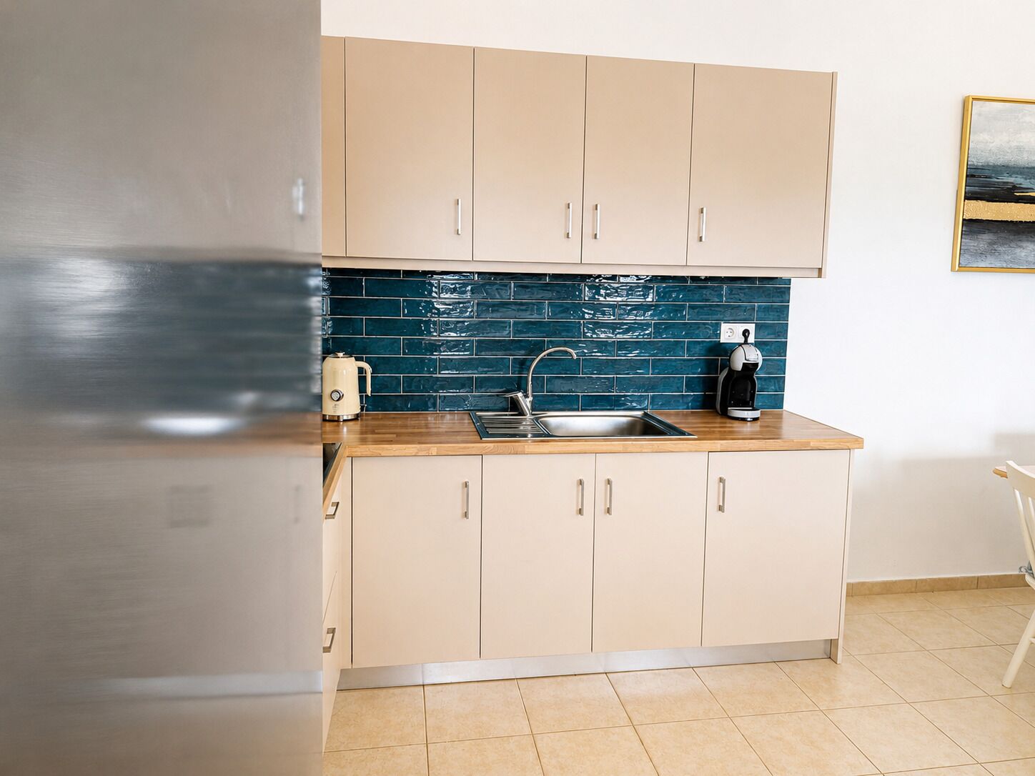 Una cucina con mobili beige, uno schienale blu, un lavandino in acciaio inossidabile e una macchina per il caffè.