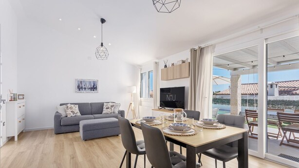 A modern living room with a sofa, dining table, and chairs. There is a TV and a window with curtains. - Villa Aurora with private pool (Tertenia)