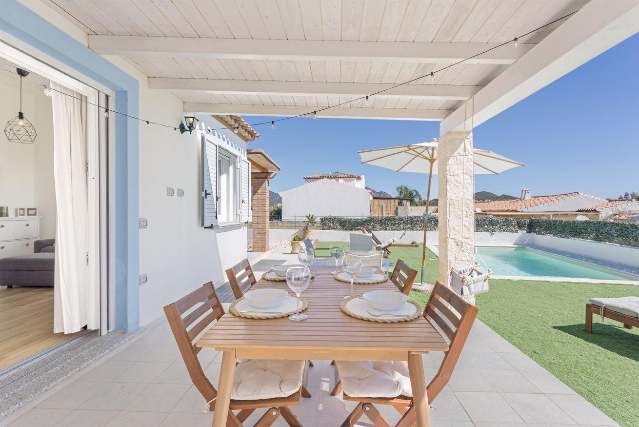 An outdoor dining area with a wooden table set for four, surrounded by chairs, and overlooking a swimming pool.