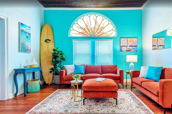 Een kamer met een blauwgroene wand, een groot raam met luiken, een surfplank, een rode bank, een klein rond tafeltje en een potplant.