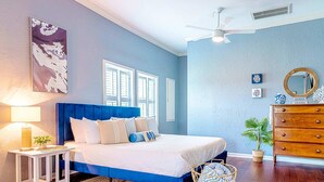 A bedroom with a large bed, a nightstand, a dresser, a ceiling fan, and a window with shutters.