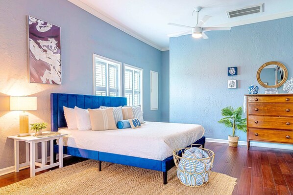 A bedroom with a large bed, a nightstand, a dresser, a ceiling fan, and a window with shutters. - Surf + Strand Dual-unit Stay (Galveston)