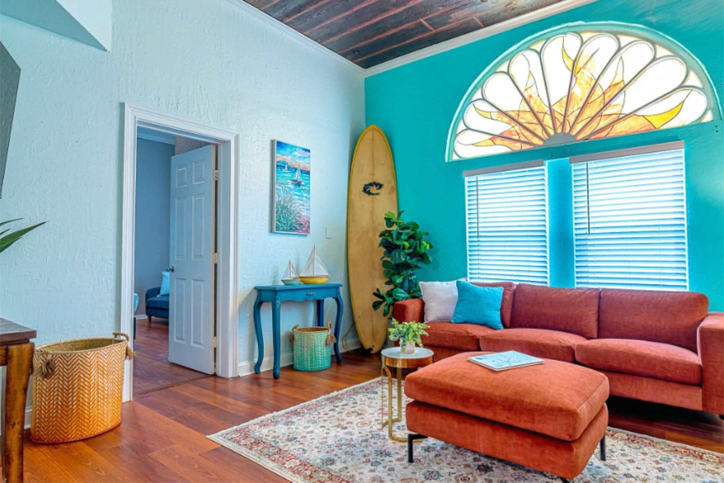 A living room with a teal wall, a wooden surfboard, a red sofa, and a window with blinds.