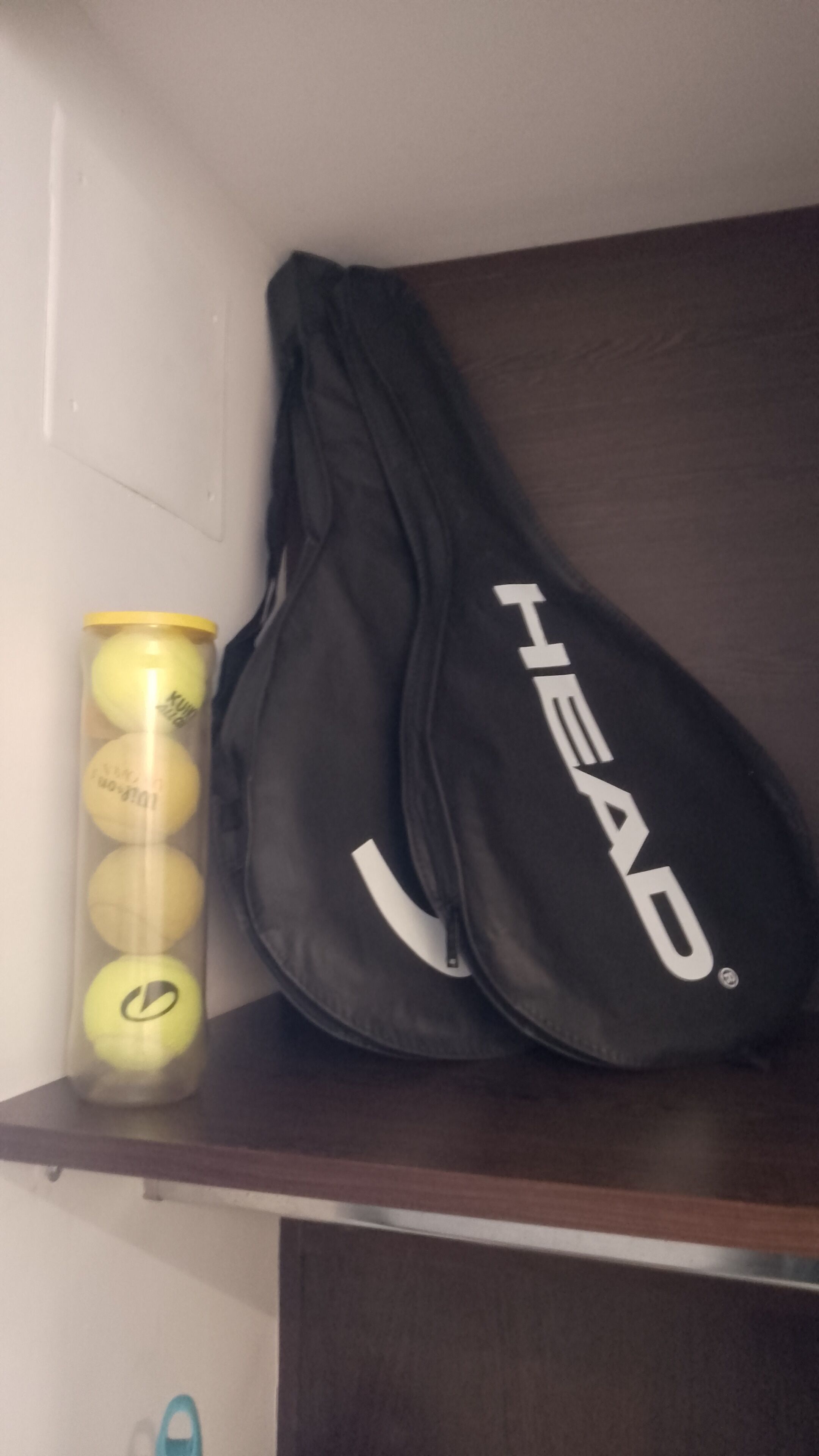 Bolsa de tenis con el nombre de la marca "HEAD" sobre una estantería junto a un recipiente con pelotas de tenis.
