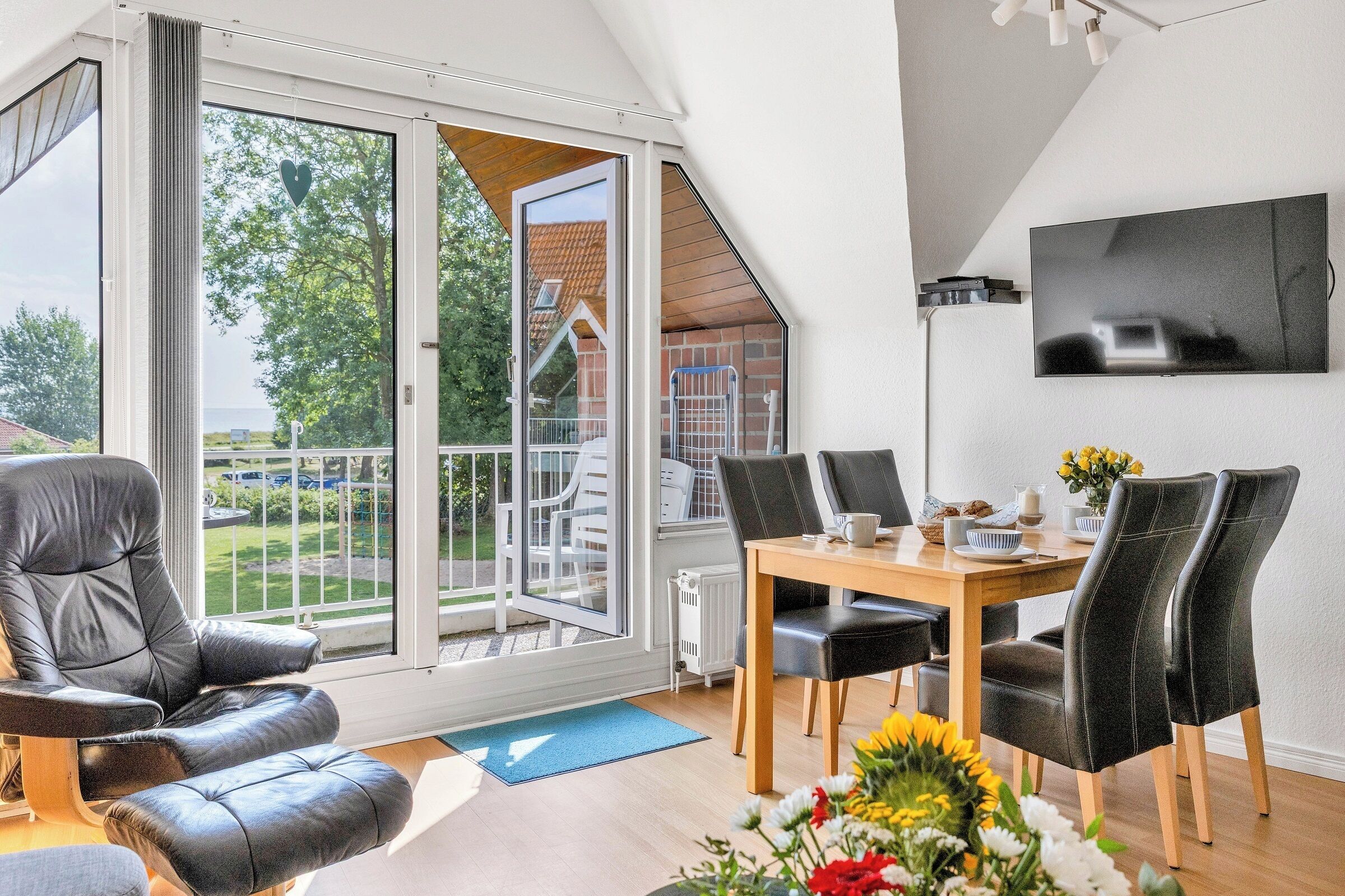 Sala de estar moderna com uma grande porta de vidro deslizante para uma varanda, área de jantar com mesa e cadeiras de madeira e uma TV de tela plana na parede.