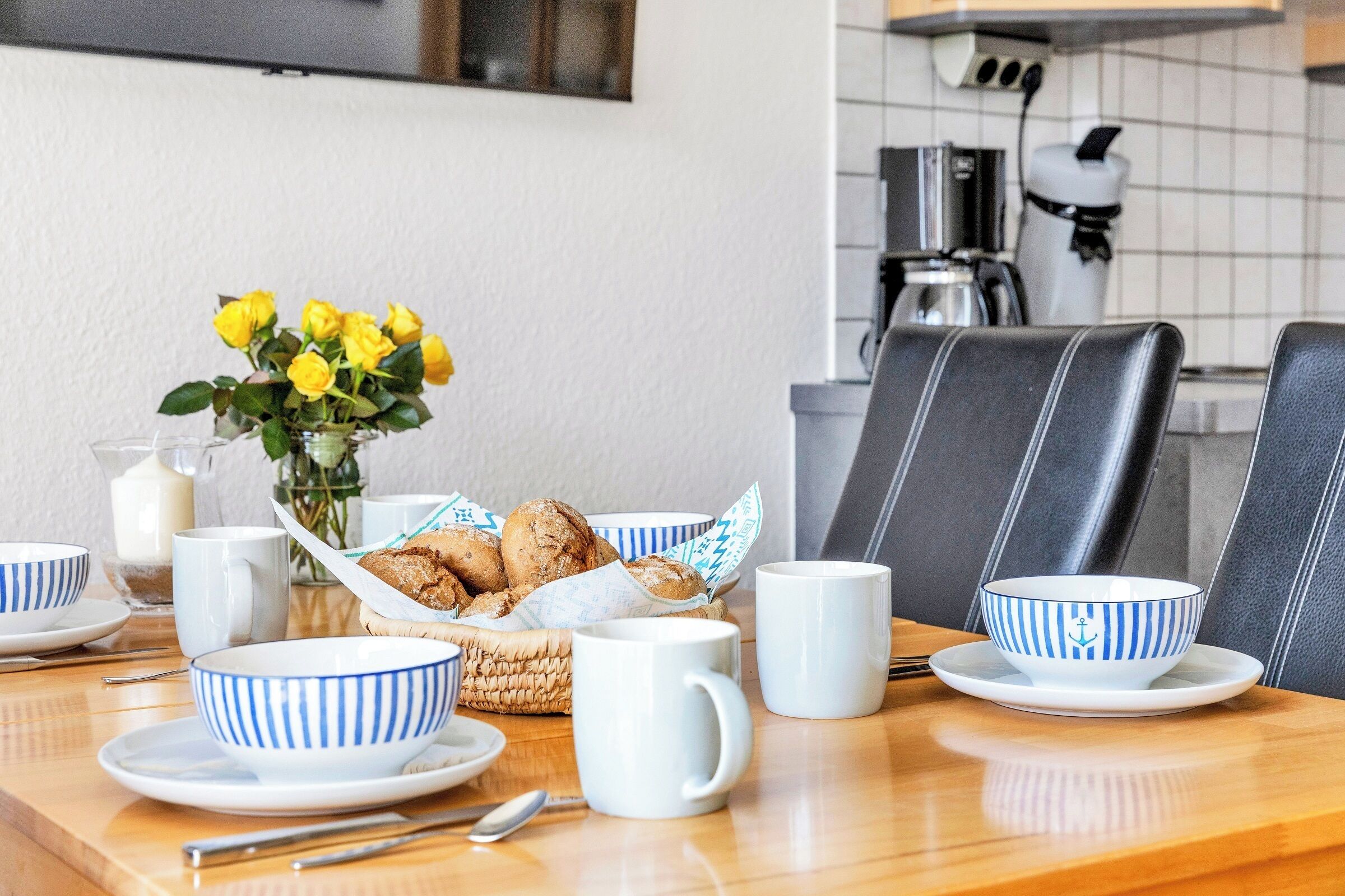 A breakfast setting with a table set for two, including a basket of pastries, a vase of yellow flowers, and matching blue and white striped bowls and cups.
