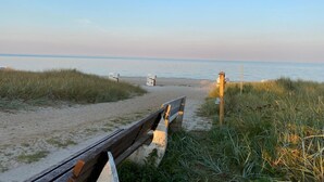 Ein Sandweg am Strand führt zum Meer, begleitet von einem Holzzaun und Strandhütten mit der Aufschrift '103'.