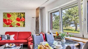 Ein modernes Wohnzimmer mit einem roten Sofa, blauen Sesseln, einem Glastisch und einem großen Fenster mit Blick auf Grünflächen.