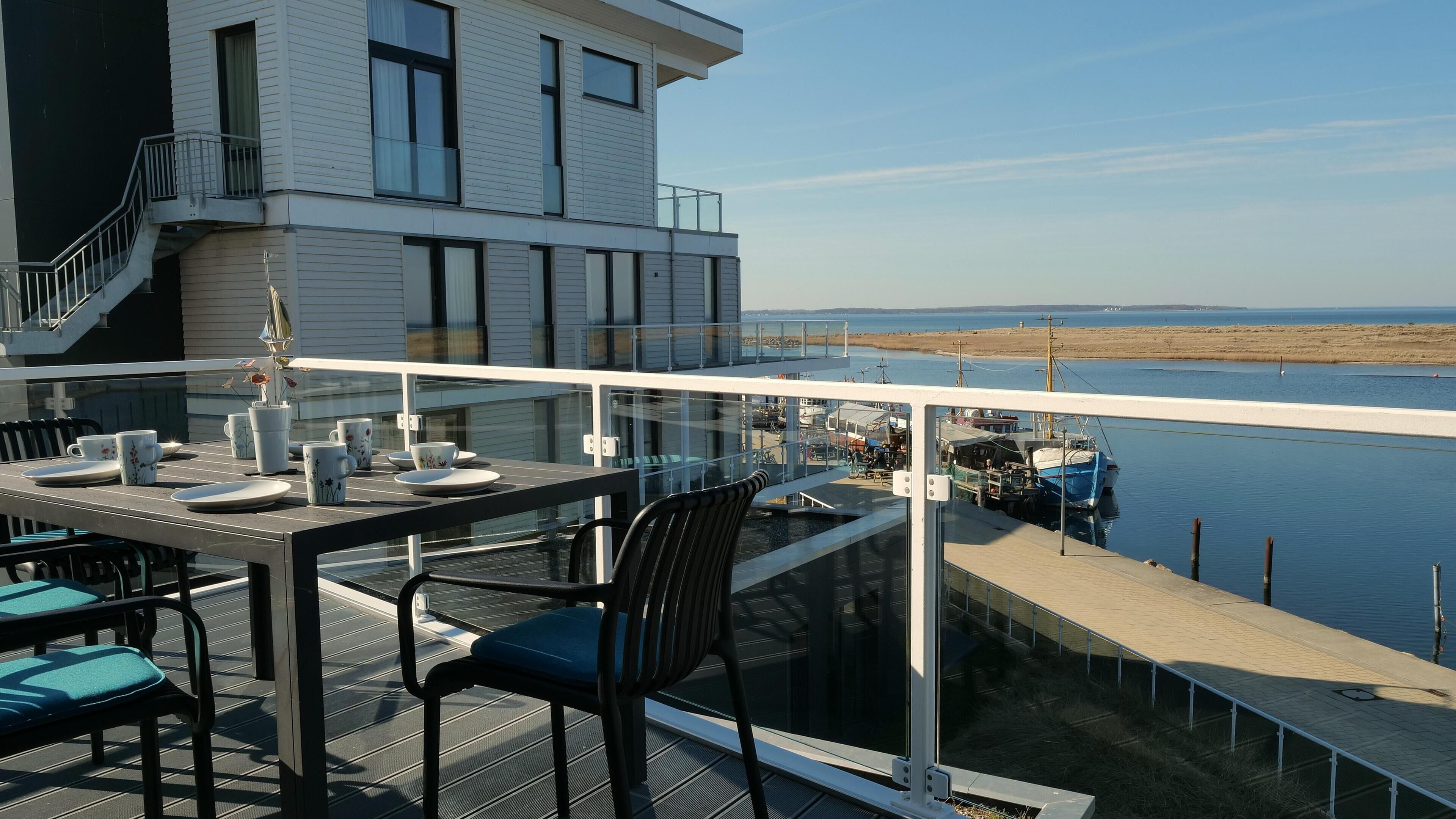 Un balcon avec une table dressée pour le dîner, donnant sur une étendue d’eau où l’on aperçoit des bateaux et une côte lointaine.