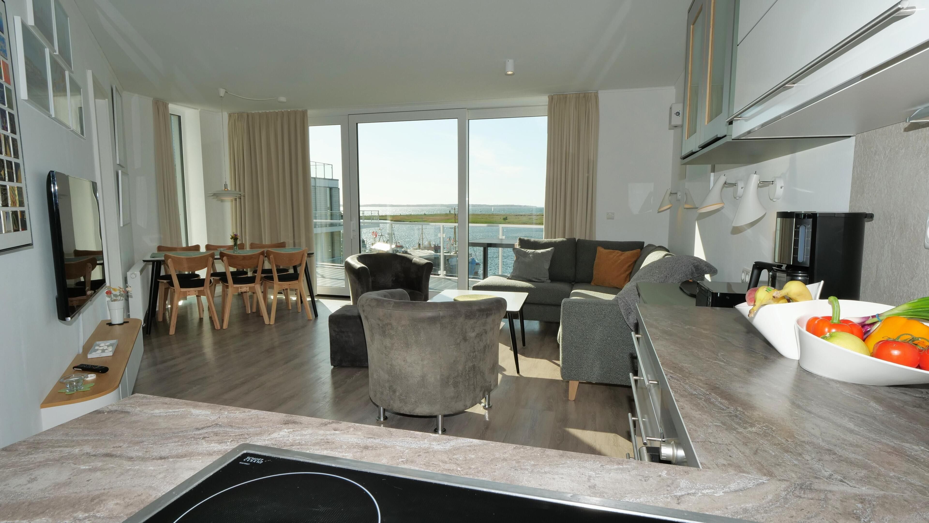 A modern living room with a sofa, chairs, a dining area, and a kitchen with a sink and fruit bowl.