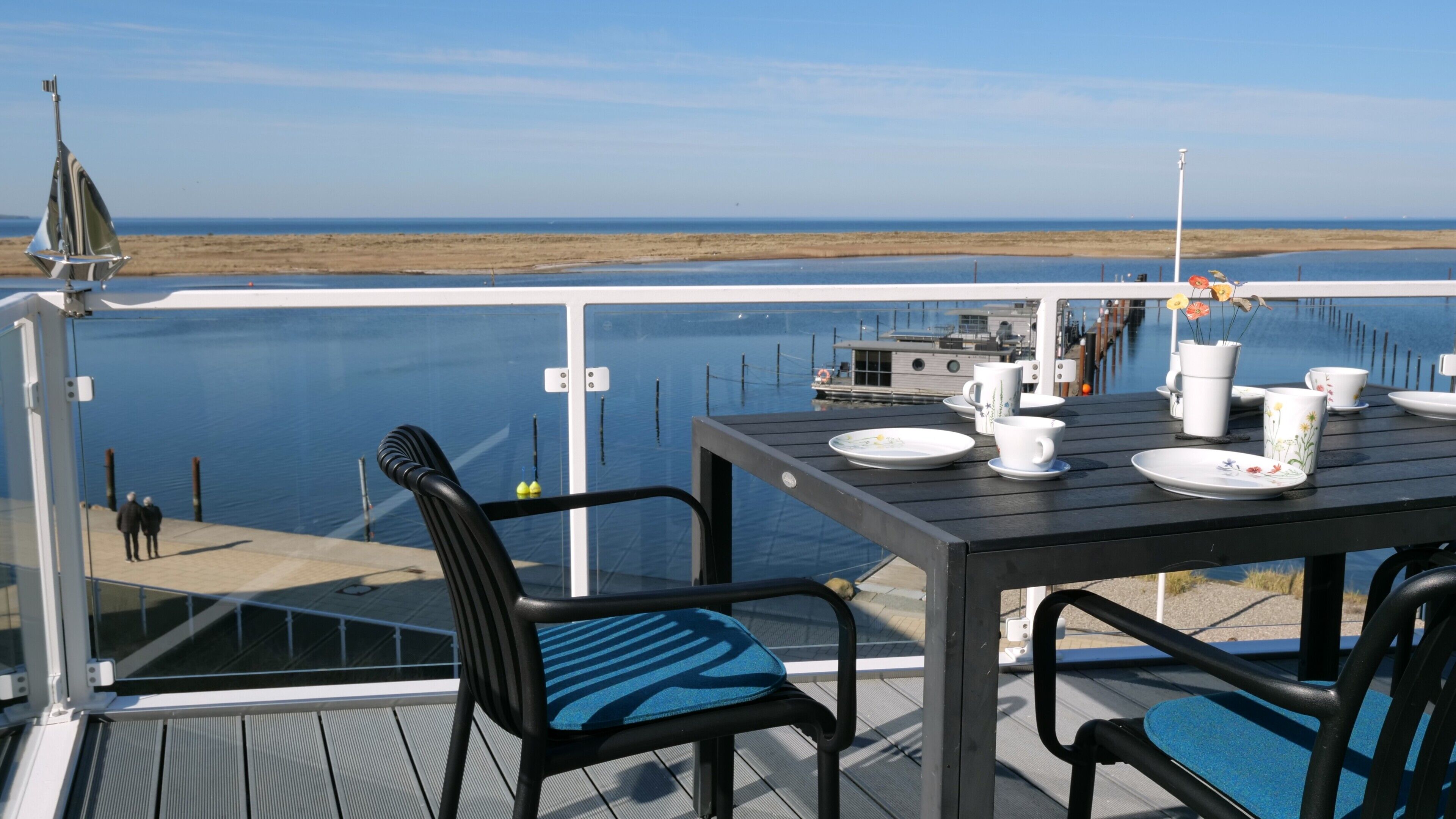 A deck with a table set for dining, overlooking a calm body of water and a distant shoreline.