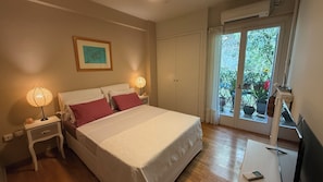 Quarto com cama, duas luminárias de cabeceira, mesa de cabeceira, rack para TV e janela com vista para o verde.