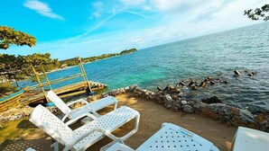 Un cadre idyllique au bord de la plage, avec des chaises longues en plastique blanc et une table, une digue rocheuse et une mer calme sous un ciel dégagé.