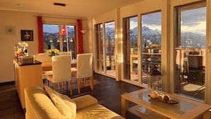 A modern living room with a sofa, dining table, and chairs, large sliding glass doors leading to a balcony with a mountain view. - swissmountainview - sunny & quiet - terrace, Jacuzzi, view to lake + mountains (Beatenberg)