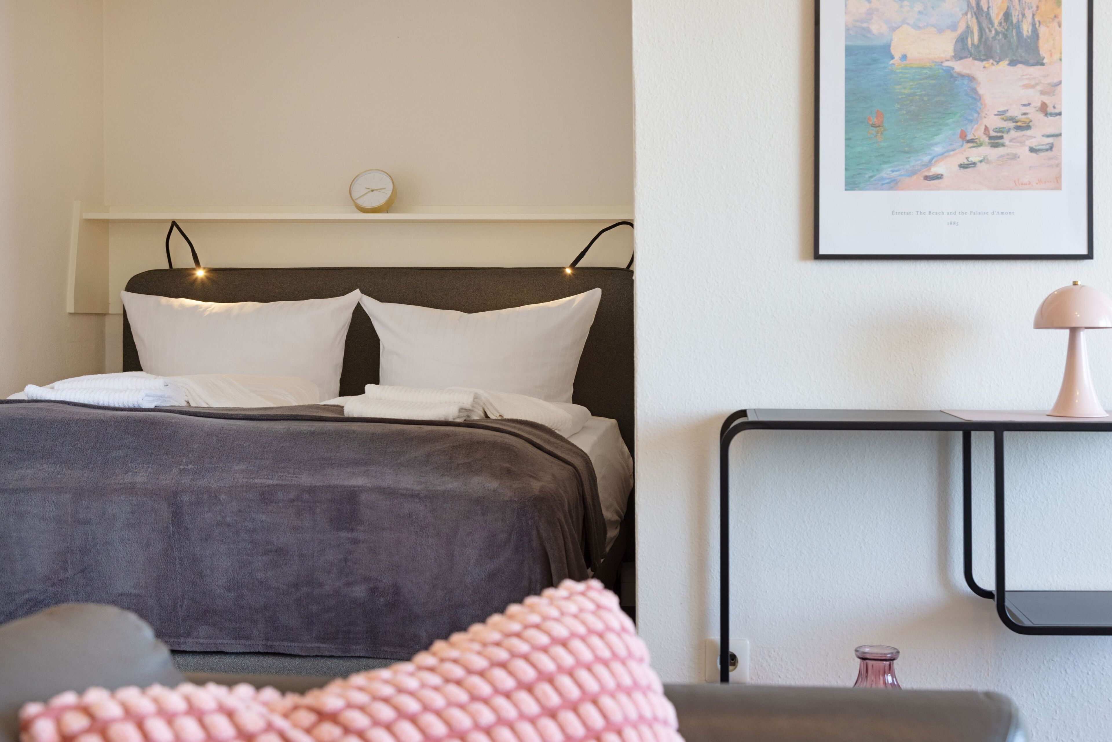 Een netjes opgemaakt bed met hoofdbord, een nachtkastje met lamp en een ingelijst schilderij aan de muur.