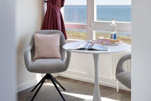 Ein Zimmer mit Meerblick, ein Tisch mit Buch und Gläsern sowie ein Sessel mit Kissen.