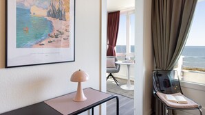 A room with a framed painting of a beach, a table with a lamp, a chair with a book, and a view of the sea through a window. - Welcome to the Best View apartment (List auf Sylt)