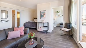 A modern hotel room with a sofa, a coffee table with flowers, a bed, a TV, and a kitchen area visible through an open door. - Welcome to the Best View apartment (List auf Sylt)