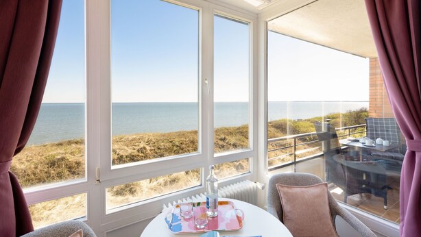 A room with a large window overlooking a beach, a table with a tray, and two chairs. - Welcome to the Best View apartment (List auf Sylt)