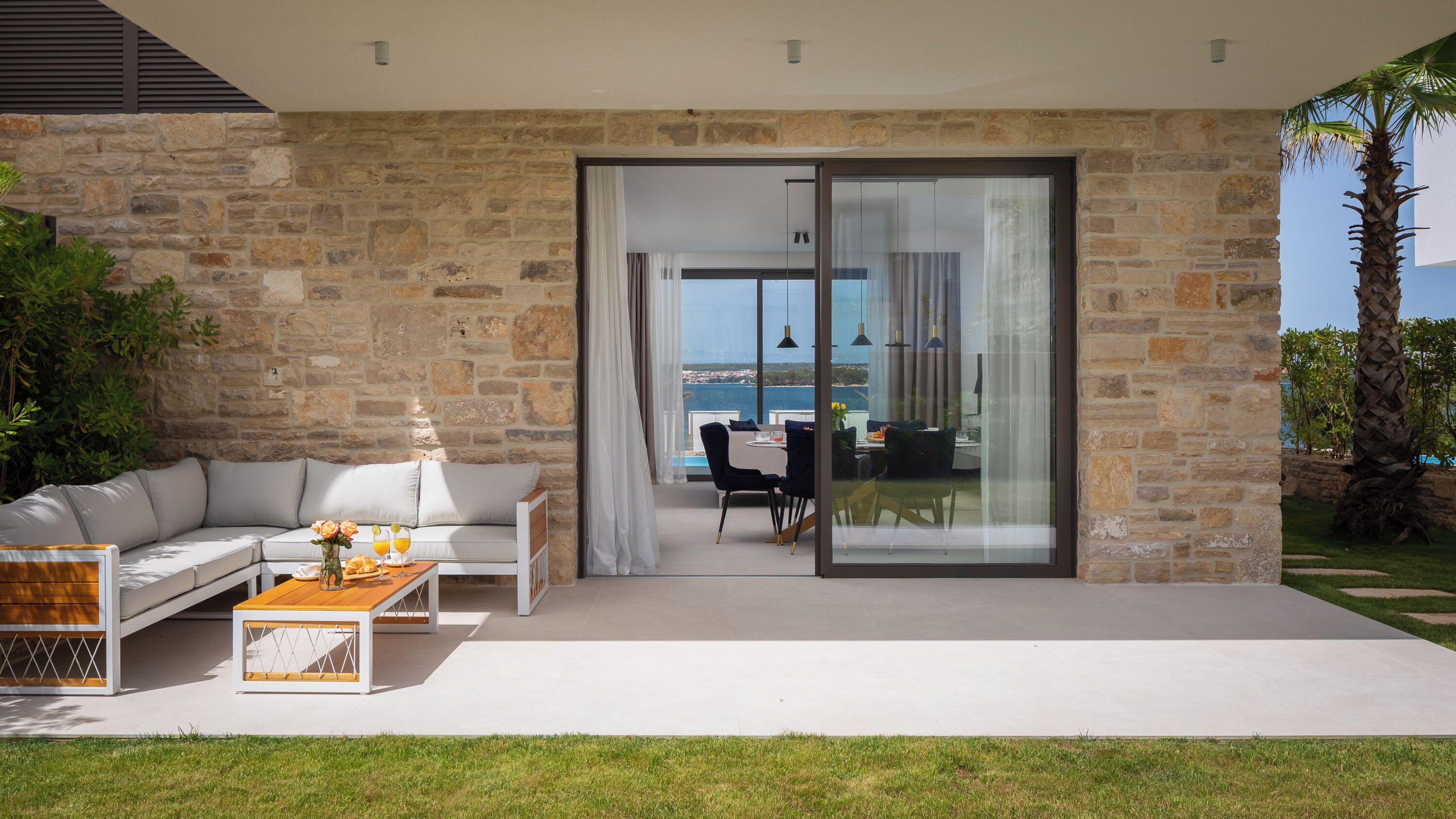 A modern outdoor living area with a stone wall, a white sofa, a dining table, and a view of the sea.