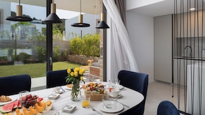 A dining area with a table set for breakfast, a vase with flowers, and a view of the garden through large windows. - The Palms Resort Villa Beach Six with Pool (Tkon)