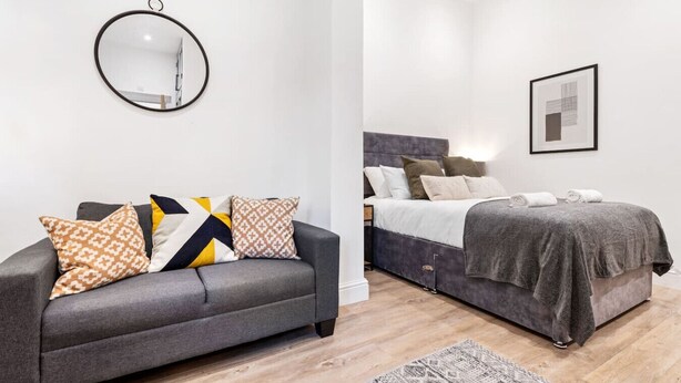 Quarto moderno com sofá cinzento, almofadas estampadas, espelho redondo, cama com cabeceira cinzenta e quadro na parede.