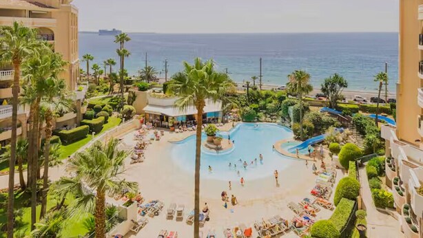 Un resort con piscina central, palmeras y vista al mar.