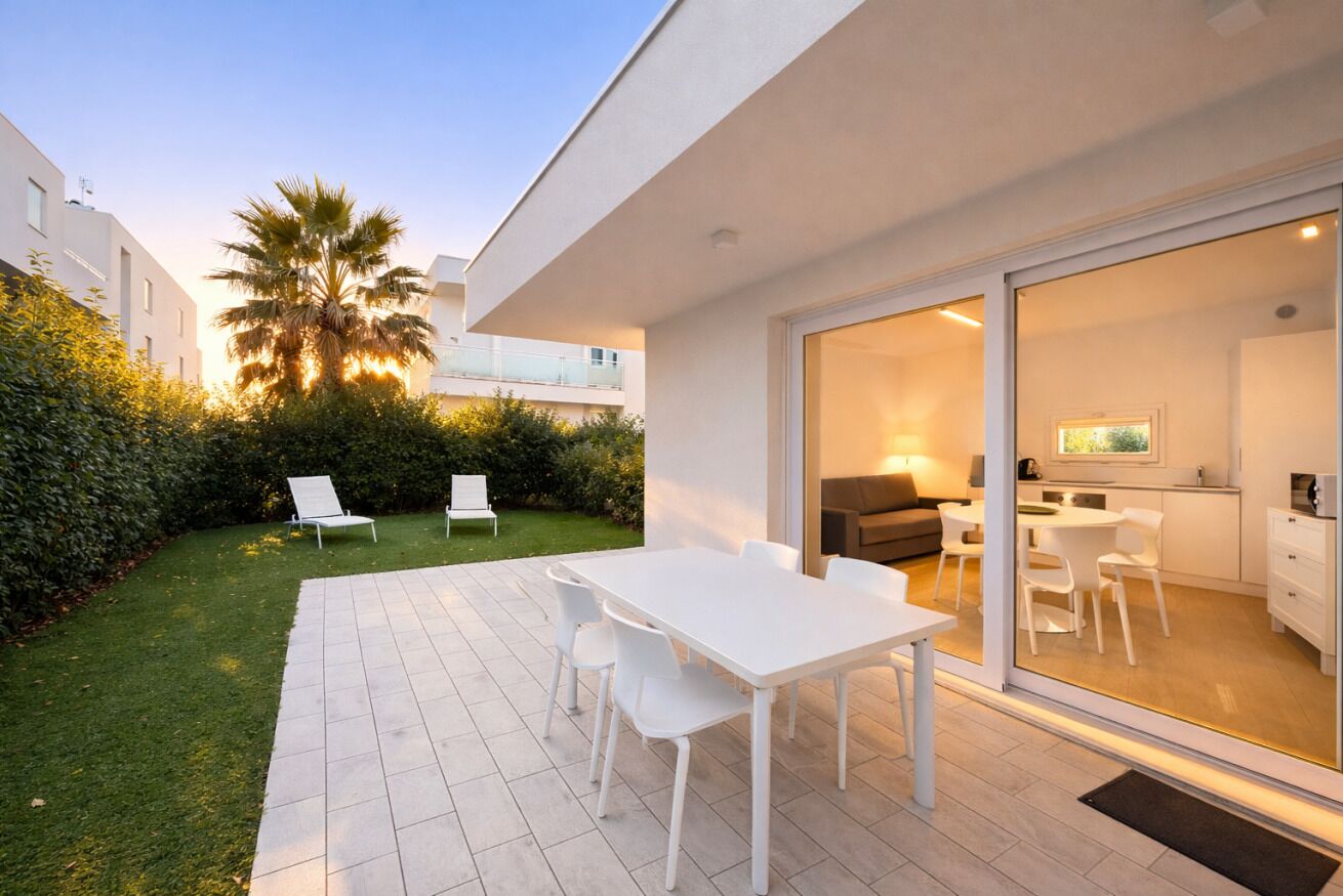 A modern outdoor patio with a white table and chairs, a grassy area, and a view of a residential building with balconies.