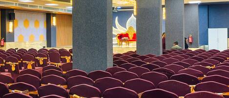 Un auditorium avec des rangées de chaises violettes, une scène avec un canapé rouge et un grand emblème doré sur le mur.