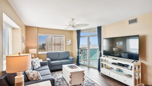 Un salon moderne avec un canapé gris, une table basse blanche, un téléviseur posé sur un meuble blanc et un balcon offrant une vue sur la ville.