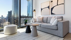 Ein modernes Wohnzimmer mit grauem Sofa, rundem Ottomanen und zwei Beistelltischen, großem Fenster mit Stadtblick und abstrakter Wandkunst.