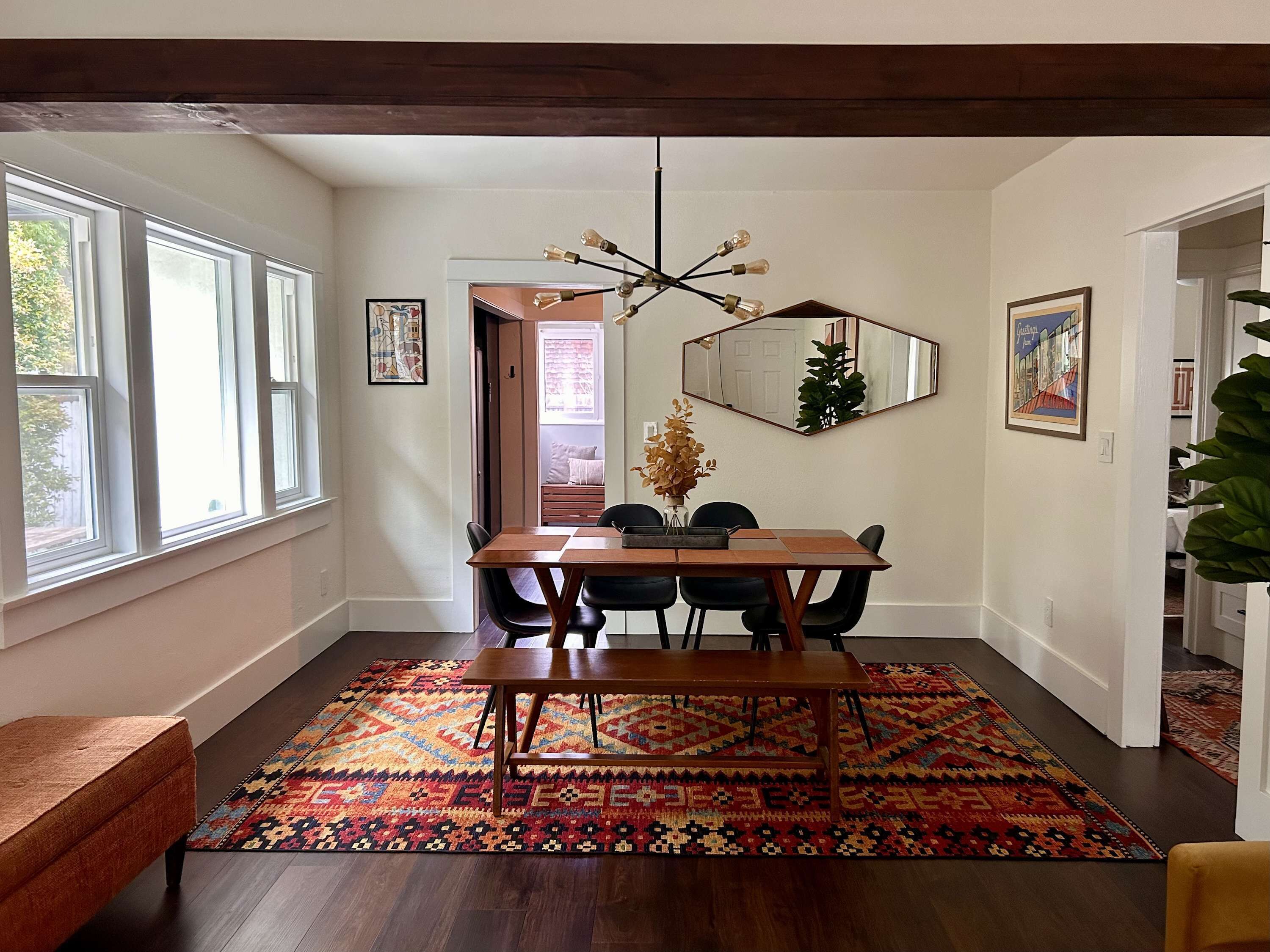 A dining area with a wooden table, black chairs, a colorful rug, a modern chandelier, and a large mirror on the wall.
