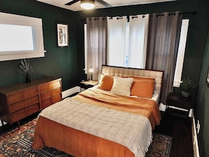 Een slaapkamer met een bed, nachtkastje, kaptafel en een raam met gordijnen.