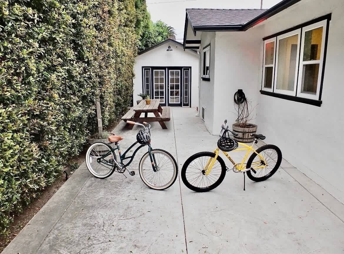 A narrow pathway leading to a white building with a black-framed window and a door. Two bicycles are parked on the pathway, one with a brown seat and the other with a yellow frame.