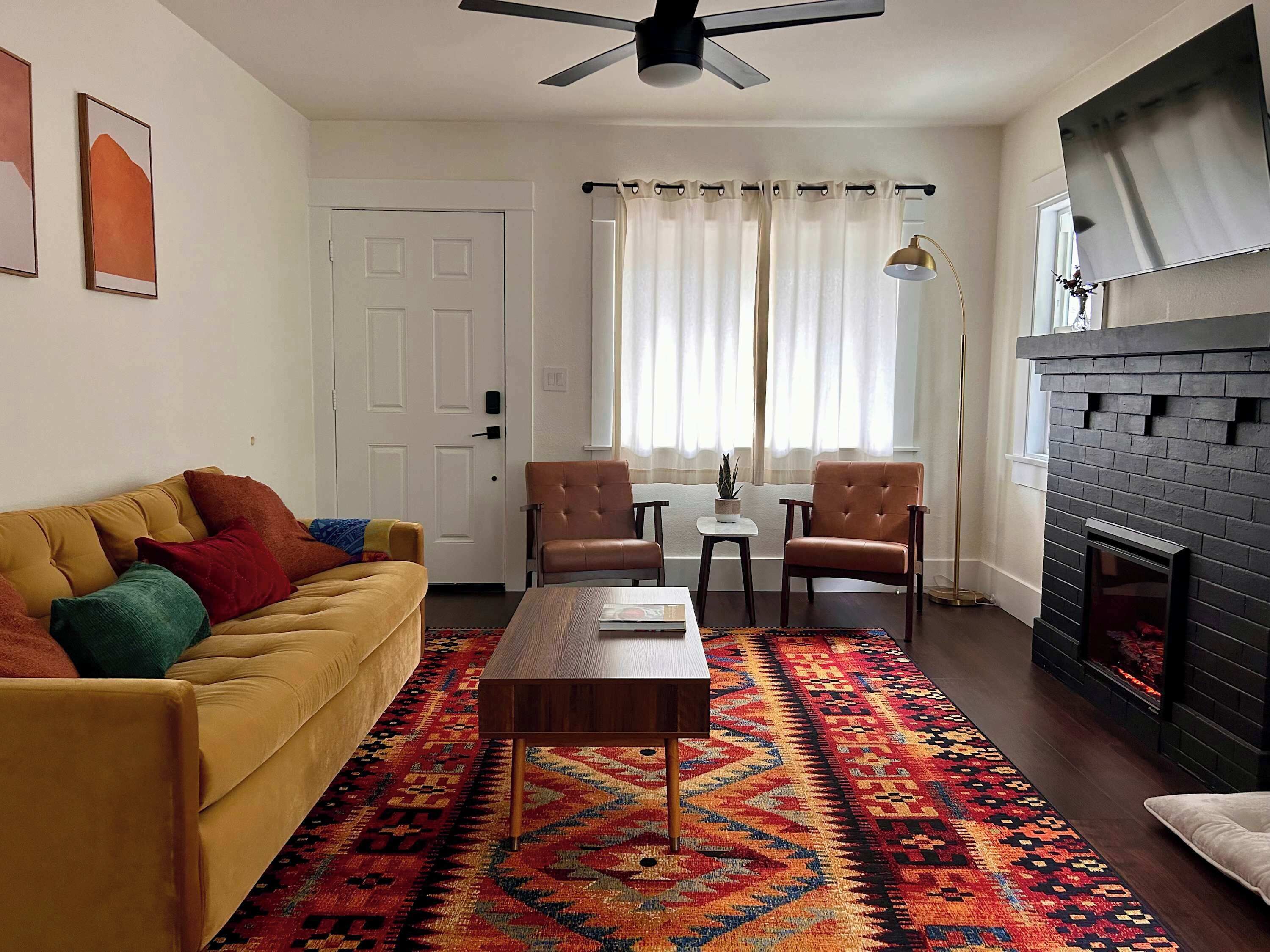 A living room with a yellow sofa, two armchairs, a coffee table, a patterned rug, a fireplace, and a ceiling fan.