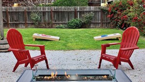 Två röda Adirondack-stolar placerade mittemot en eldstad med glasbord, där en eld brinner.
