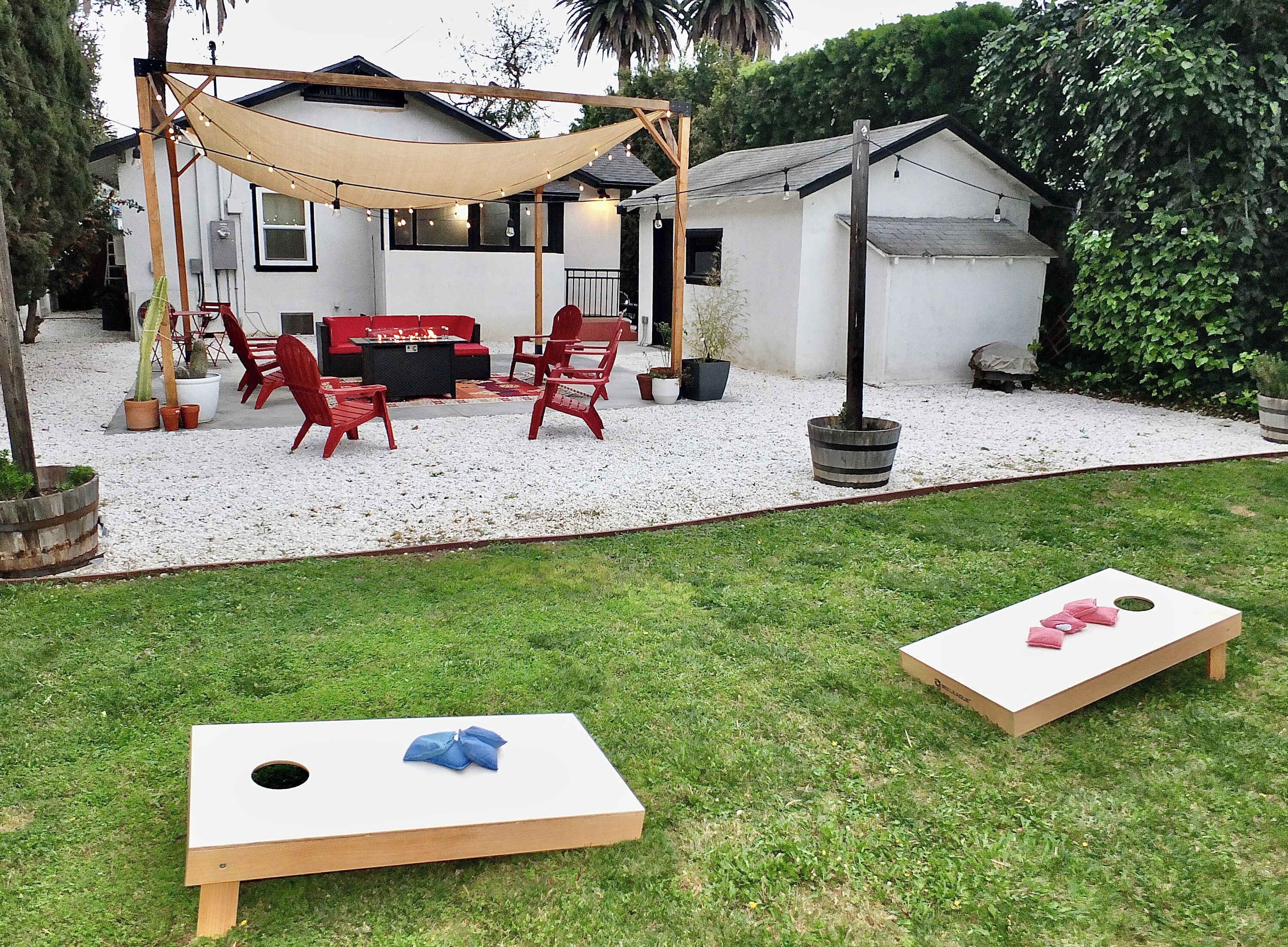 Un patio trasero con zona de barbacoa, sillas rojas y un toldo. Hay dos juegos de lanzamiento de maíz en el césped.