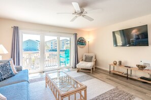 A living room with a sofa, a glass coffee table, a chair, a TV, and a balcony with a view of buildings. - Joanie's Gem - 1st Floor Surf Condo - Sleeps 6! (Surf City)