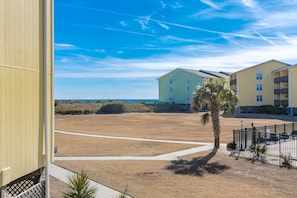 A building with a flat roof and a balcony, situated next to a sandy area with a palm tree. - Joanie's Gem - 1st Floor Surf Condo - Sleeps 6! (Surf City)